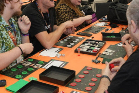 Group of people playing a board game at a table with various game pieces and boxes.